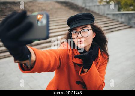 Jeune femme en manteau et chapeau prenant des photos d'elle-même sur un téléphone portable dans la rue Banque D'Images