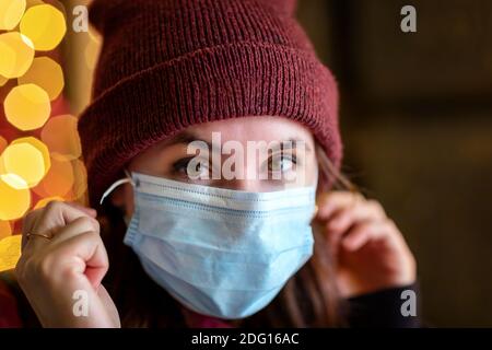 Jeune fille avec masque de protection et chapeau de laine regarde les fenêtres du magasin décorées avec des lumières de Noël. Banque D'Images