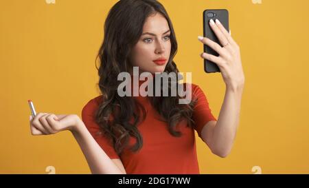 Belle fille brunette en toute confiance appliquant rouge rouge rouge à lèvres regardant dans smartphone sur fond jaune Banque D'Images