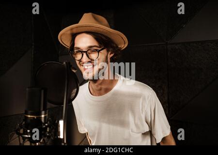 Beau chanteur souriant chantant volontiers en train d'enregistrer une nouvelle chanson pour un album de musique dans un studio moderne. Musicien attrayant travaillant en studio d'enregistrement Banque D'Images