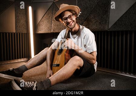 Jeune homme élégant et attrayant avec guitare, heureux de regarder dans l'appareil photo assis au sol dans un studio d'enregistrement Banque D'Images