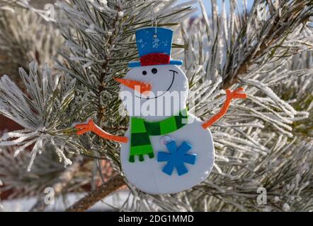Gros plan d'un bonhomme de neige en peluche dans un chapeau et une écharpe accrochés sur le sapin de Noël à l'extérieur pendant la saison hivernale. Branches de sapin recouvertes de neige. Hiver Banque D'Images