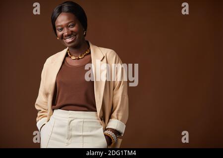 Portrait à la taille haute d'une élégante femme afro-américaine debout sur fond marron en studio et souriant à l'appareil photo, espace de copie Banque D'Images