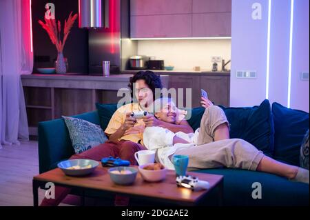 Joyeux petit ami et petite amie riant après avoir joué à des jeux vidéo à la maison, des joysticks et des collations sur une table. Couple du millénaire appréciant passer du temps Banque D'Images