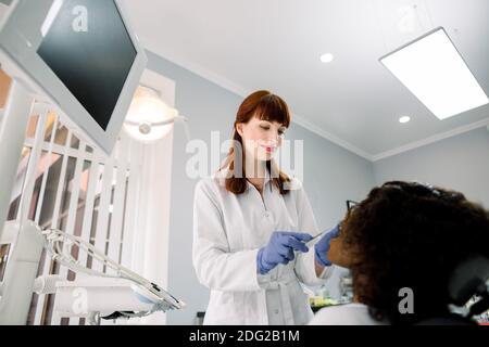 Jolie femme dentiste portant des gants, effectuant des vérifications dentaires et des traitements sur les dents des patients. Femme afro-américaine couchée à la chaise, recevant Banque D'Images