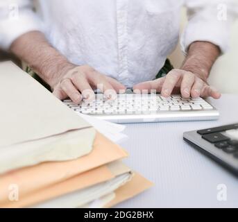 Les mains d'un homme travaillant sur son clavier d'ordinateur. Il est entouré de documents administratifs et d'une calculatrice. Banque D'Images