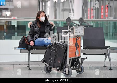 Voyages à thème en période de pandémie de corona. Jeune femme avec masque, masque avec sa valise, bagages à l'aéroport de Munich le 6 décembre 2020. Assis dans le terminal, hall des départs, en attente du départ. MODÈLE DISPONIBLE ! | utilisation dans le monde entier Banque D'Images