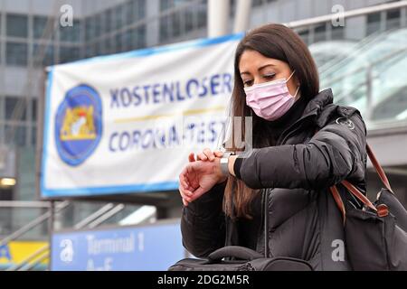 Voyages à thème en période de pandémie de corona. Jeune femme avec masque, masque à l'aéroport de Munich le 6 décembre 2020. Elle sort du terminal avec ses bagages et sa valise, derrière elle se trouve un panneau D'ESSAI GRATUIT CORONA. Elle regarde sa montre. MODÈLE DISPONIBLE ! | utilisation dans le monde entier Banque D'Images
