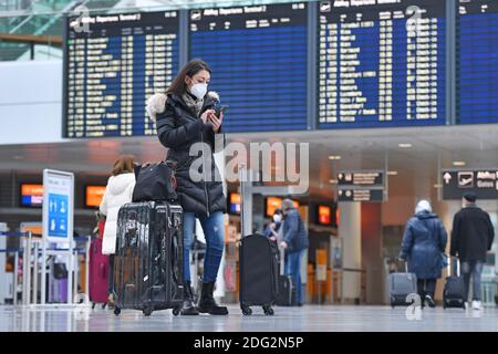 Voyages à thème en période de pandémie de corona. Jeune femme avec masque, masque avec sa valise, bagages à l'aéroport de Munich le 6 décembre 2020. Regarde son smartphone - derrière elle se trouve le tableau des départs. MODÈLE DISPONIBLE ! | utilisation dans le monde entier Banque D'Images