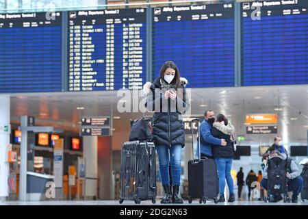 Voyages à thème en période de pandémie de corona. Jeune femme avec masque, masque avec sa valise, bagages à l'aéroport de Munich le 6 décembre 2020. Regarde son smartphone - derrière elle se trouve le tableau des départs. MODÈLE DISPONIBLE ! | utilisation dans le monde entier Banque D'Images