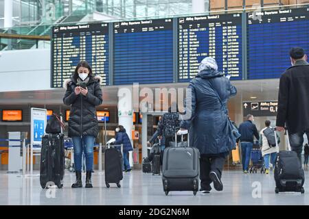 Voyages à thème en période de pandémie de corona. Jeune femme avec masque, masque avec sa valise, bagages à l'aéroport de Munich le 6 décembre 2020. Regarde son smartphone - derrière elle se trouve le tableau des départs. MODÈLE DISPONIBLE ! | utilisation dans le monde entier Banque D'Images