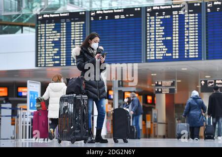 Voyages à thème en période de pandémie de corona. Jeune femme avec masque, masque avec sa valise, bagages à l'aéroport de Munich le 6 décembre 2020. Regarde son smartphone - derrière elle se trouve le tableau des départs. MODÈLE DISPONIBLE ! | utilisation dans le monde entier Banque D'Images