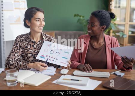 Portrait de deux sourires de deux femmes d'affaires regardant chacune autre et l'utilisation de l'ordinateur tout en étant assis à la table dans le bureau contre le mur vert Banque D'Images