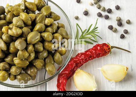Gros plan de câpres marinées dans une soucoupe en verre, de la gousse de piment rouge, de l'ail et des grains de poivre noir sur une table rustique en bois. Bourgeons marinés de caper bush. Banque D'Images