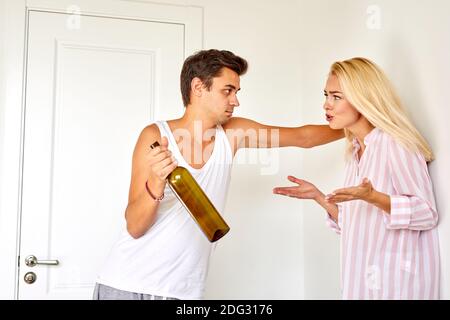 la femme crie avec colère à son mari ivre à la maison, la femme blonde blâme le mari alcoolique. l'alcoolisme dans la famille. Un gars avec une bouteille dans son ha Banque D'Images