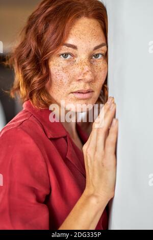 femme à l'estime en robe rouge regarde la caméra penchée sur le mur blanc en studio, portrait Banque D'Images