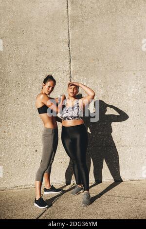 Pleine longueur de femme et son entraîneur de fitness debout à l'extérieur contre un mur un matin ensoleillé. Les femmes en forme de sport portent debout ensemble regardant Banque D'Images