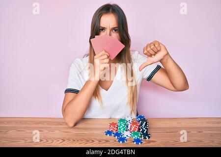 Belle femme caucasienne jouant au poker de jeu couvrant le visage avec des cartes avec le visage en colère, signe négatif montrant dislike avec les pouces vers le bas, rejet c Banque D'Images