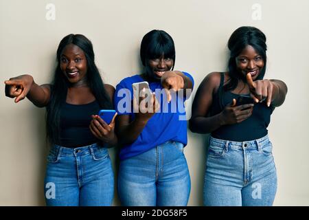 Trois jeunes afro-américains amis utilisant un smartphone pointant vers vous et l'appareil photo avec les doigts, souriant positif et gai Banque D'Images