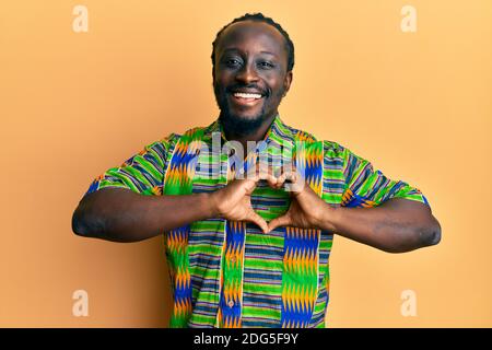 Beau jeune homme noir portant des vêtements ethniques colorés souriant dans l'amour faisant le symbole de coeur forme avec les mains. Concept romantique. Banque D'Images