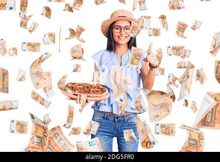 Jeune belle fille latine portant un chapeau d'été et des lunettes tenant pizza souriante et positive, pouce vers le haut faisant excellent et signe d'approbation Banque D'Images