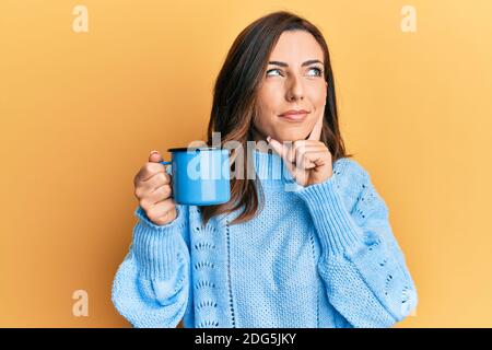 Jeune femme brune tenant une tasse de café sérieux visage pensant à la question avec la main sur le menton, réfléchi à la confusion idée Banque D'Images