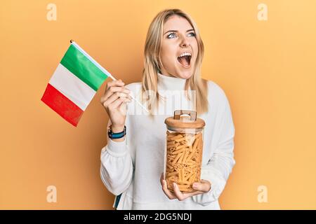 Jeune femme caucasienne tenant un pot de pâtes macaroni et drapeau italien en colère et fou hurlant frustré et furieux, criant avec colère. Rage et agg Banque D'Images