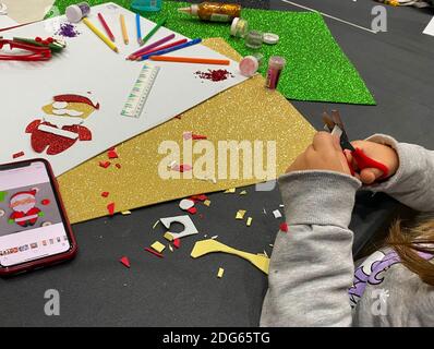 Gros plan des mains d'une fille en utilisant les ciseaux pour faire des métiers de Noël avec du carton coloré. Téléphone portable sur la table. Banque D'Images