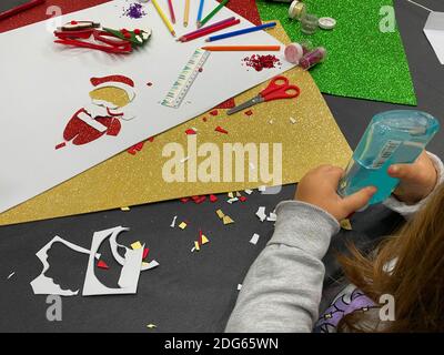 Gros plan des mains d'une fille en utilisant la colle pour faire un métier de Noël. Paillettes, crayons de couleur, ciseaux, carton de couleur et règle éparpillés sur le Banque D'Images