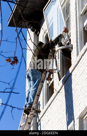 Un homme de réparation hispanique portant des jeans et des baskets se trouve au-dessus d'une échelle de construction qui remplace la fenêtre d'une maison de briques vintage. Un collègue est serviable Banque D'Images