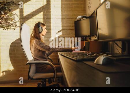 Jeune femme dans un casque ayant des appels vidéo par ordinateur à la maison. Travail à distance, lumière du soleil sur le mur. Écran vide pour votre photo. Banque D'Images