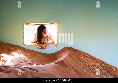 Jeune jolie femme accrochée à la fenêtre de la maison abandonnée au milieu du désert, brunette portant des lunettes de soleil, maison pleine de sable ondulé Banque D'Images