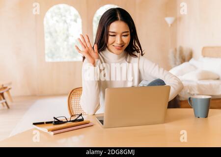 Une jeune femme asiatique porte un pull avec un joli sourire en agitant quelqu'un dans un ordinateur portable dans une chambre confortable. Des conférences et des appels vidéo pour filles heureux et élégants. Portrait d'affaires d'hiver style de vie. Copier l'espace Banque D'Images