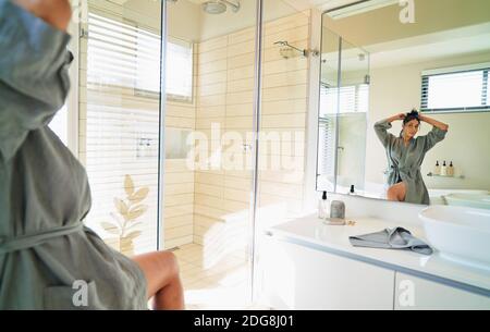 Femme en peignoir fixant les cheveux dans le miroir de salle de bains ensoleillé Banque D'Images