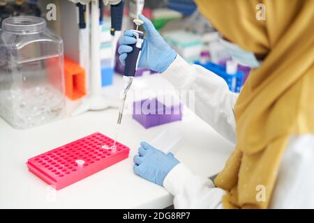 Scientifique féminine dans le hijab et le masque facial remplissant le plateau de pipettes en laboratoire Banque D'Images