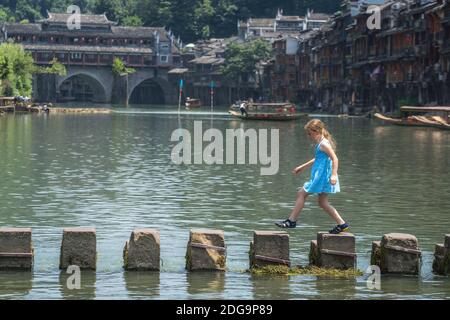 Une jeune fille caucasienne traverse des eaux sur des pierres sur la rivière Tuojiang, qui traverse le centre de la vieille ville de Fenghuang, en Chine Banque D'Images