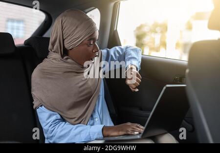 Étant en retard. Femme d'affaires musulmane noire stressée, assise dans un taxi et à l'heure de vérification Banque D'Images