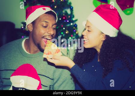 Couple de manger de la pizza pour la nourriture de noël et de s'amuser. Un beau jeune couple qui mange de la pizza, qui parle et qui souriait à la maison. Arbre de Noël sur fond. Banque D'Images