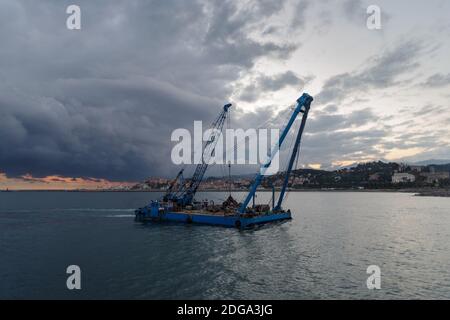 Une barge de grue de construction flotte le long de la côte pendant les travaux de remise en état des terres au large de la côte de Ligurie, en Italie Banque D'Images