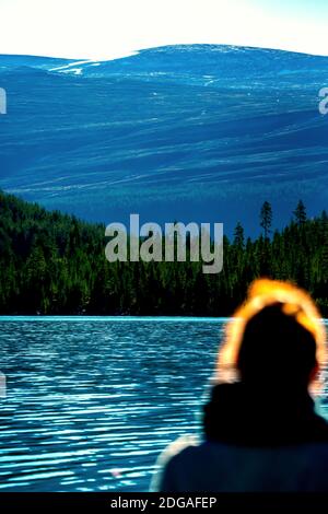 Silhouette floue d'une femme debout près d'un grand lac dans la nature surplombant la forêt de pins verdoyants et les montagnes enneigées. Premier plan flou. Banque D'Images