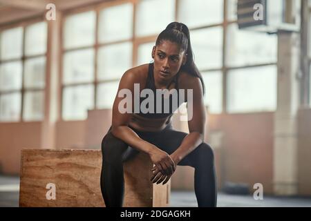 Femme ethnique musclée dans les vêtements de sport assis sur le cube et regardant loin pendant la pause dans l'entraînement fonctionnel dans la salle de sport spacieuse Banque D'Images