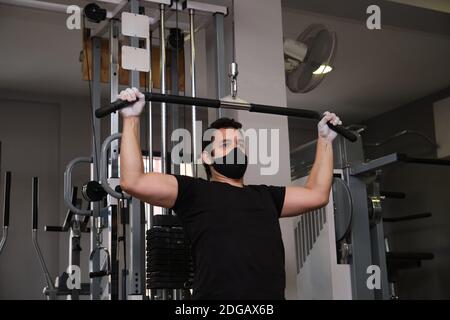 Homme sportif portant un masque de protection pour faire de l'exercice avec une machine à tirer lat pour construire le muscle de bras et d'épaule dans la salle de fitness. Carrosserie nouvelle norme Banque D'Images