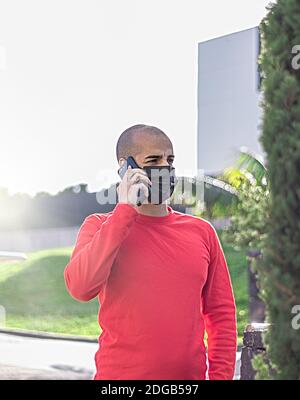 Jeune homme avec masque de contrôle de son téléphone mobile et parlant dans le parc Banque D'Images