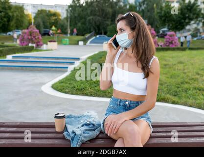 une femme en été dans la ville appelle sur un smartphone, un masque médical protecteur sur son visage, la quarantaine et le verrouillage en été dans la ville dans la nature. En ligne Banque D'Images