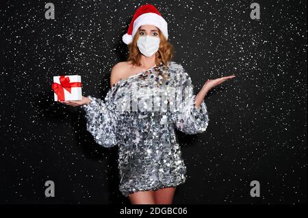 Jeune femme en robe de fête brillante, chapeau de père Noël, et masque de protection médicale avec boîte cadeau de Noël tient le produit sur la paume. Banque D'Images