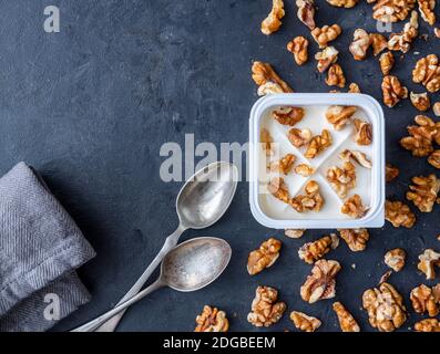 Yogourt blanc dans une tasse carrée en plastique avec noix et cuillères sur fond gris. Vue à plat. Banque D'Images