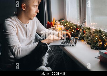 Jeune homme freelance en pajama à distance de travail en ligne ayant appel vidéo sur ordinateur portable avec chat au bureau à domicile. Ambiance de Noël, nouvel an décorati Banque D'Images