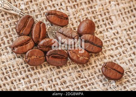 Cuillère métallique avec grains de café torréfiés aromatisés gros plan sur fond de sac de toile de jute, mise au point sélective. Concept de service et de restauration Banque D'Images