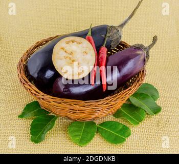 Deux aubergines matures entières avec un poivron dans un brun osier panier sur fond jaune clair Banque D'Images