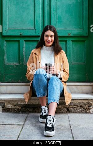 Portrait d'une jeune femme élégante assise à l'extérieur et utilisant un smartphone Banque D'Images